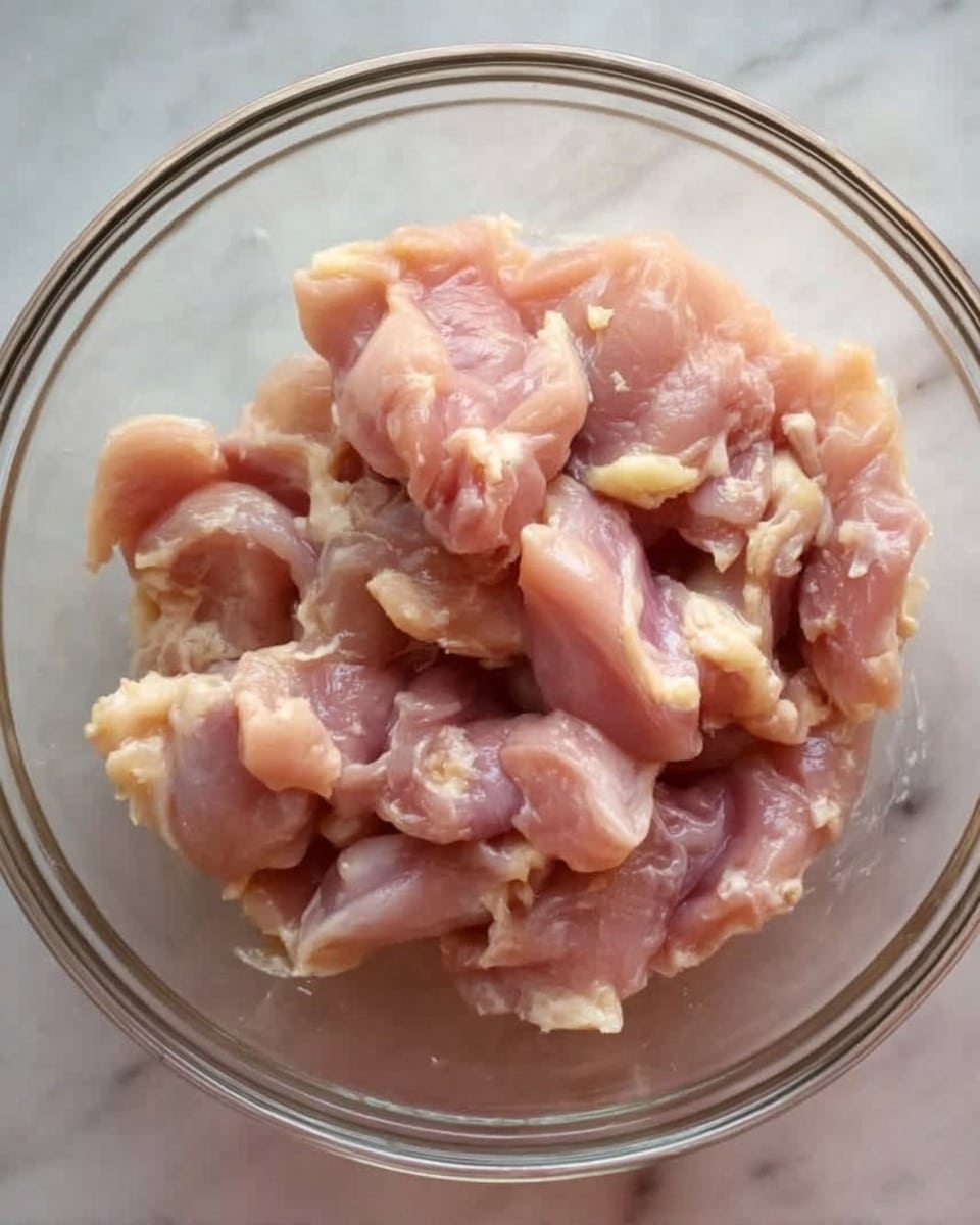 A clear glass bowl filled with raw, pale pink chicken pieces that have a soft, slightly shiny texture. The bowl is placed on a white marbled surface. The chicken pieces look fresh, with some light yellow fat spots visible, piled loosely inside the bowl. Photo taken with an iphone --ar 4:5 --v 7