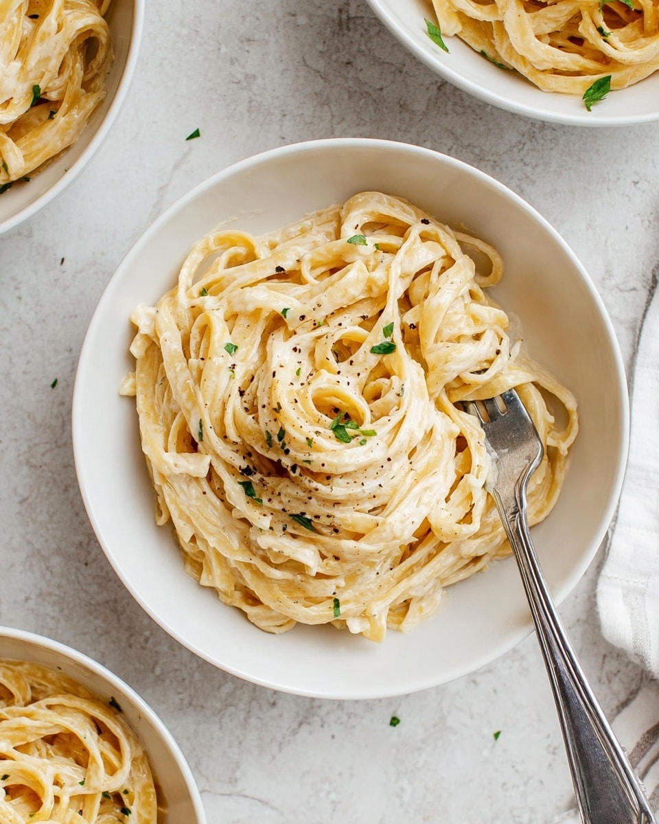 A white round bowl filled with creamy fettuccine pasta coated in a smooth, light-colored sauce, sprinkled with small green herb pieces and a few black pepper specks on top. The pasta strands are thick and twisted loosely, showing their soft texture. A silver fork rests inside the bowl, with some pasta twirled around its tines. The bowl is set on a white marbled surface, and parts of two more white bowls with the same pasta are visible around the main bowl. photo taken with an iphone --ar 4:5 --v 7
