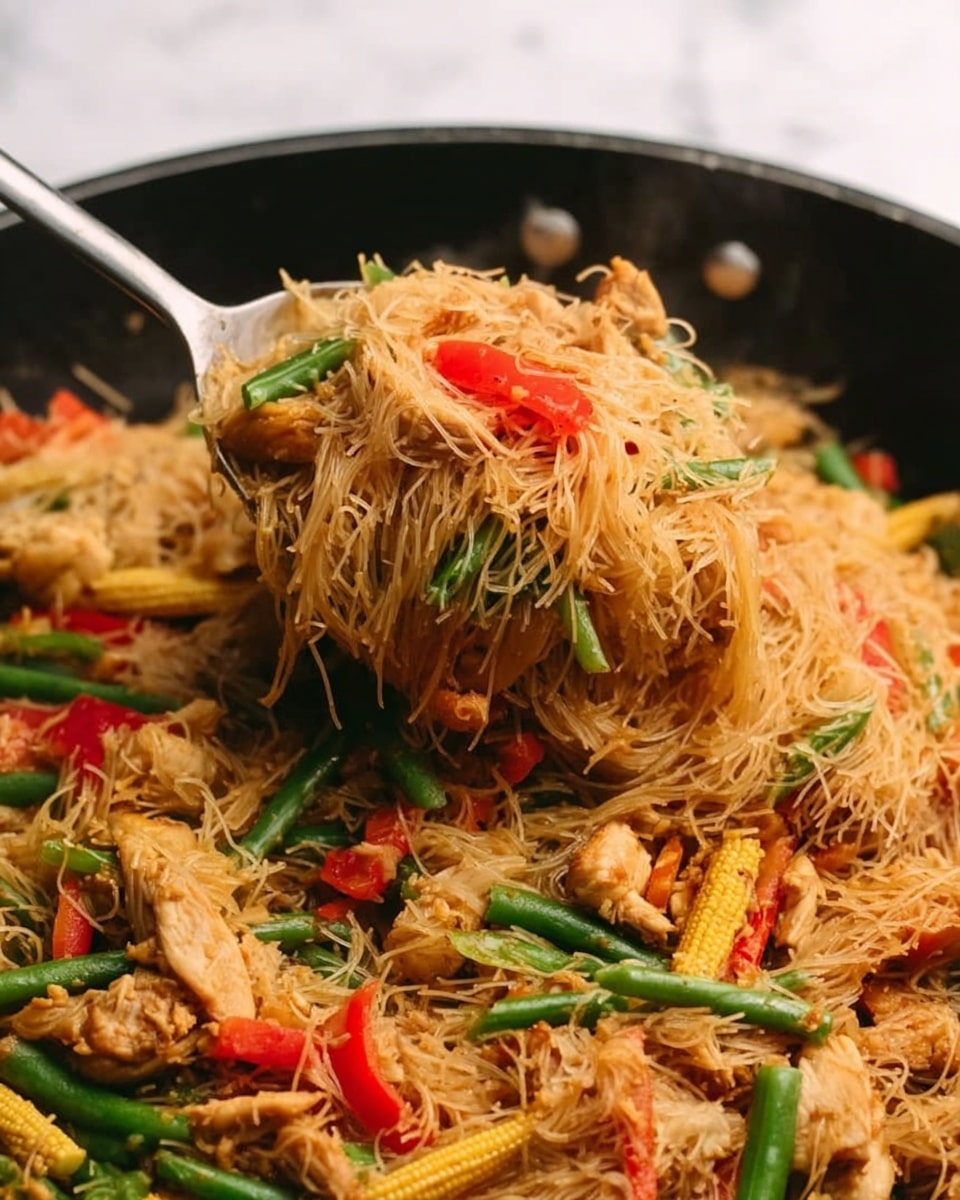 A close-up of a large black pan filled with stir-fried thin rice noodles mixed with pieces of cooked chicken, bright green beans, sliced red bell peppers, and baby corn. The noodles have a soft, slightly shiny texture, and the vegetables add pops of bright green, red, and yellow colors scattered throughout. A metal spoon is partially visible, lifting a large heap of the noodle dish, showing its tangled layers and mixed ingredients. The background is a white marbled surface. photo taken with an iphone --ar 4:5 --v 7