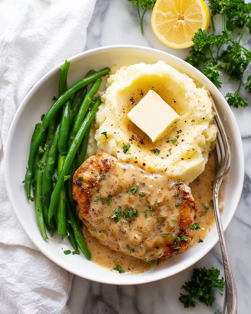 A white round plate sits on a white marbled surface with a serving of mashed potatoes, green beans, and a piece of chicken covered in creamy gravy. The mashed potatoes are soft and creamy with a square of melting butter on top, sprinkled with black pepper, positioned on the right side of the plate with a fork resting in them. At the bottom of the plate are bright green beans, shiny and neatly arranged in a row. On the left side is a grilled chicken piece, golden brown with a thick layer of rich, light brown gravy sprinkled with fresh green parsley bits. A lemon wedge peeks out from behind the chicken near the top right edge of the plate. The background shows a white cloth and some parsley leaves with a lemon slice on the white marbled texture. photo taken with an iphone --ar 4:5 --v 7