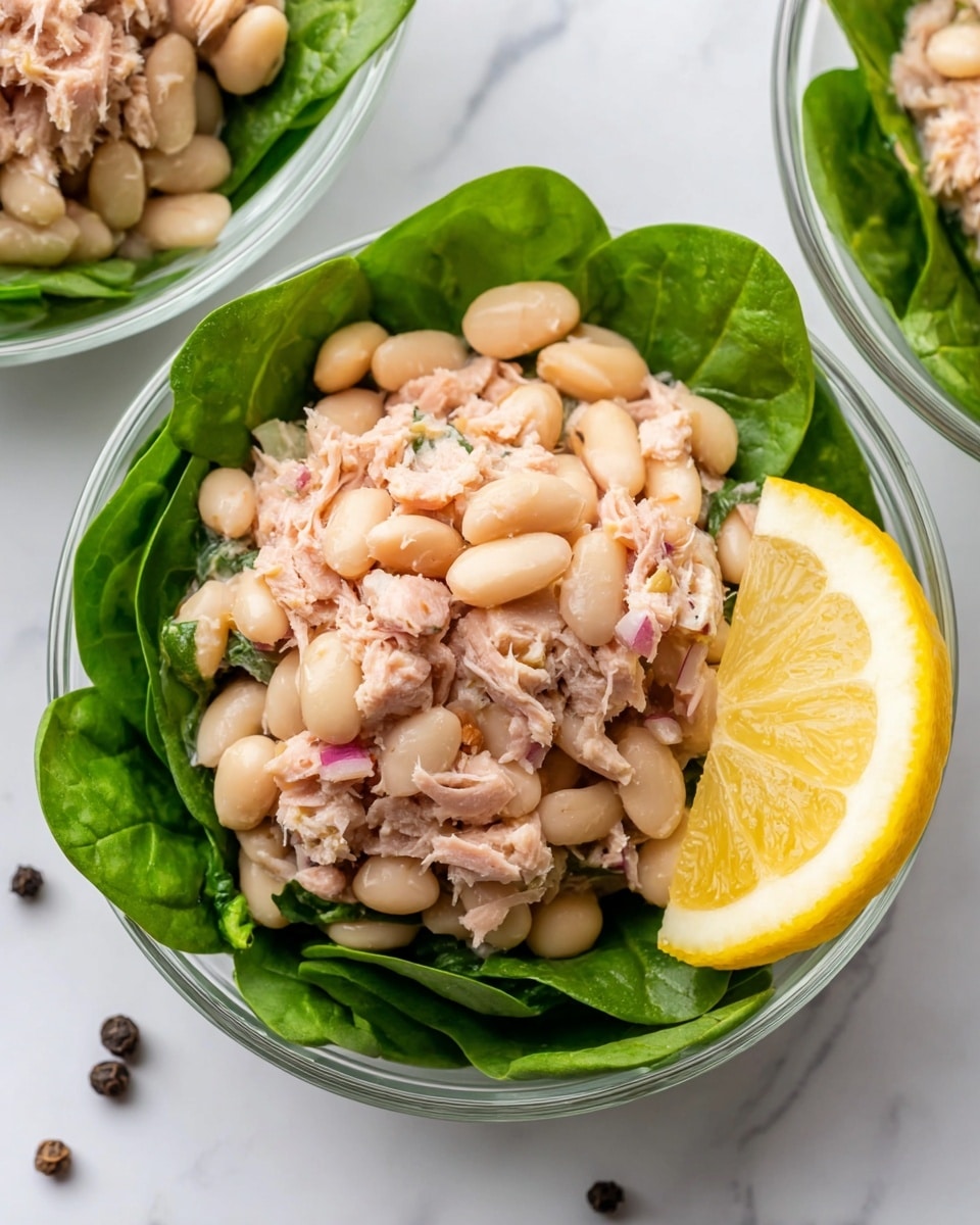 The image shows a clear glass bowl filled with a fresh tuna and white bean salad. The bottom layer is bright green spinach leaves, neatly lined to form a base. On top is a chunky mixture of light beige white beans, flaked pink tuna, and small bits of red onion, creating a textured and hearty layer. A thin slice of lemon with a vibrant yellow peel and translucent pale yellow inside leans against the salad on the right side of the bowl. The bowl sits on a white marbled surface, with a few whole black peppercorns scattered nearby. Photo taken with an iphone --ar 4:5 --v 7