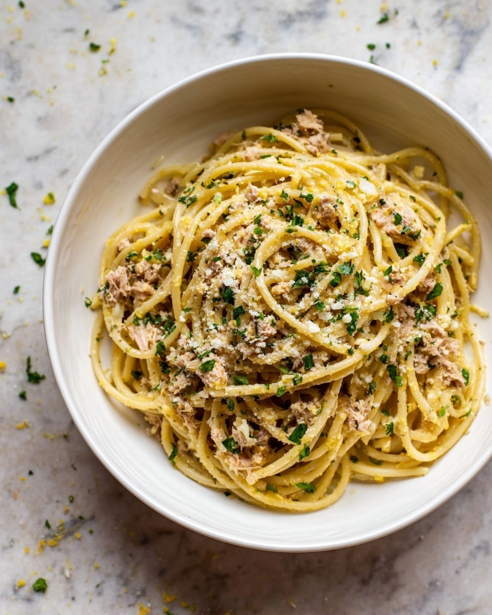 A white bowl filled with a round nest of spaghetti pasta, light yellow in color, mixed with small pieces of tuna scattered evenly throughout. The pasta is lightly coated with a glossy sauce, sprinkled with finely chopped green herbs and a light dusting of grated white cheese on top. The bowl sits on a white marbled surface with a few small bits of herbs scattered around it. Photo taken with an iphone --ar 4:5 --v 7