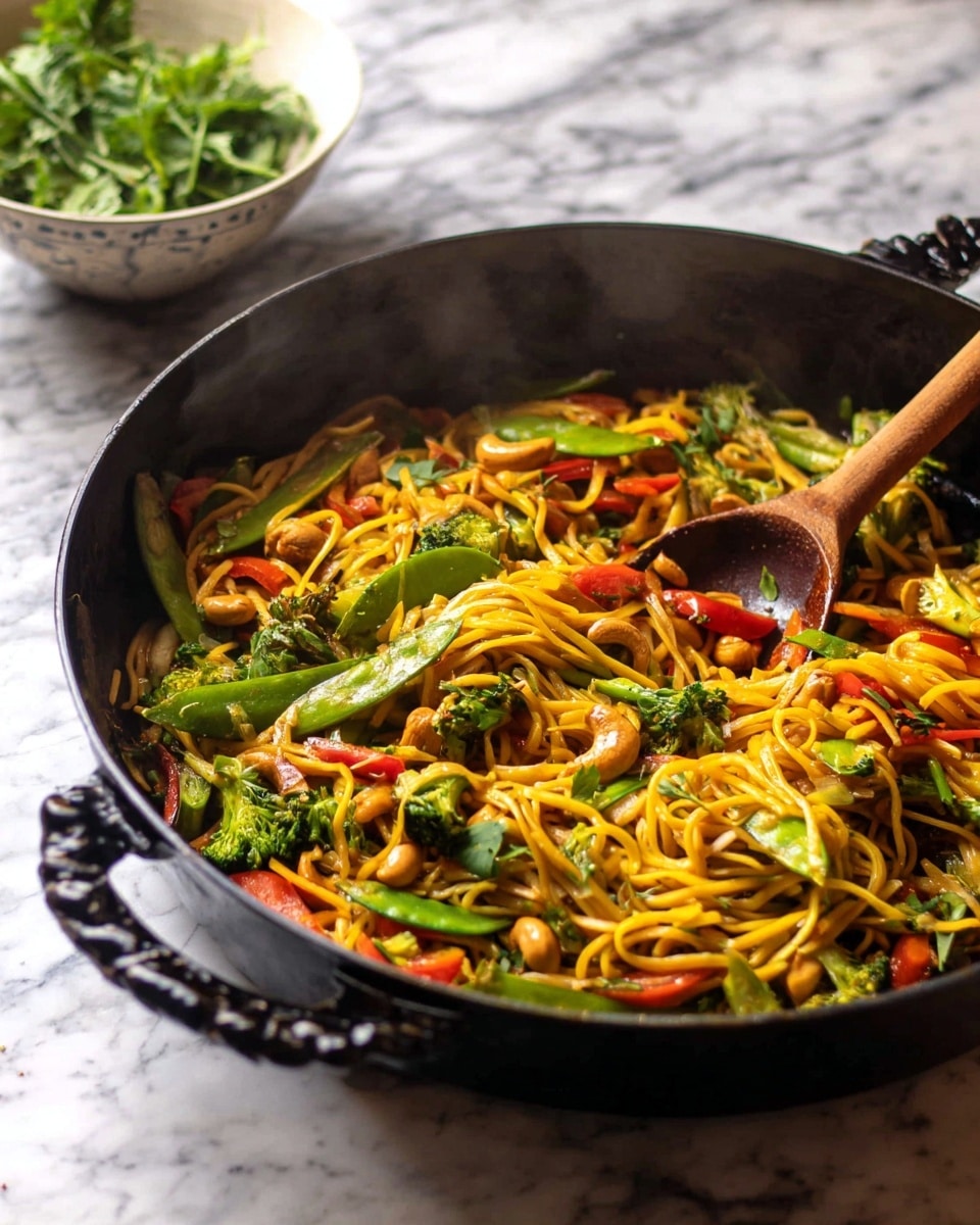 A large black cast iron pan filled with steaming stir-fried yellow noodles mixed with bright green snap peas, small pieces of broccoli, red bell pepper slices, and chunks of light brown cashews, all coated in a glossy sauce. A wooden spoon is resting inside the pan, partially covered with the noodles and vegetables. In the background, a white bowl containing fresh green herbs sits on a white marbled surface. photo taken with an iphone --ar 4:5 --v 7