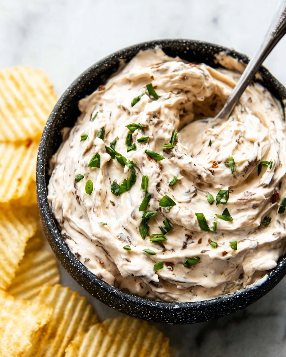 A close-up view of a black speckled bowl filled with a thick creamy dip that has a pale beige color with visible dark bits mixed throughout. The dip is swirled in layers with a rough but smooth texture and is sprinkled on top with chopped fresh green herbs, likely scallions. A metal spoon is partially dipped into the creamy mixture, resting inside the bowl. Around the bowl are some light yellow, ridged potato chips placed on a white marbled surface. photo taken with an iphone --ar 4:5 --v 7