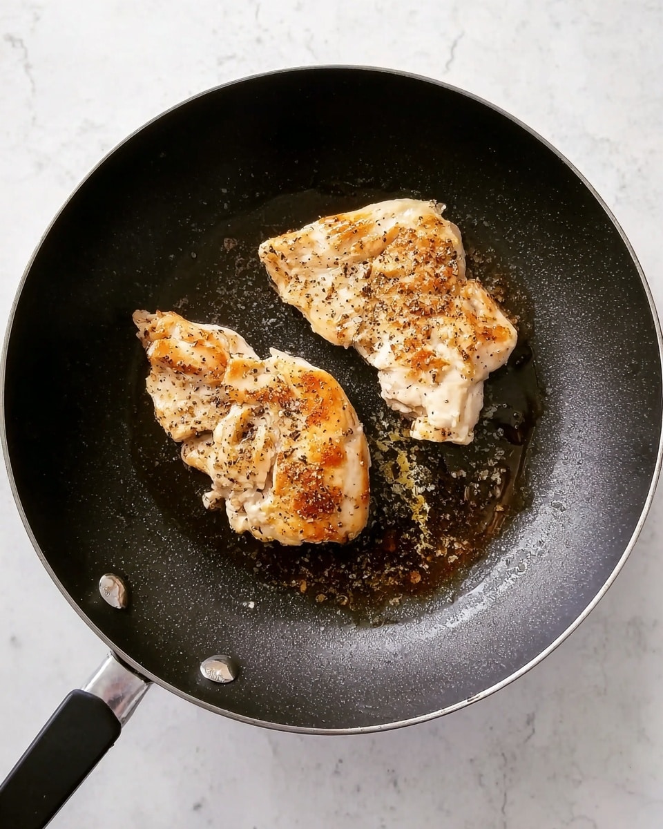 Two pieces of cooked chicken are in a dark non-stick frying pan. The chicken pieces are light brown with some black pepper and a bit of oil around them. The pan is on a white marbled surface. photo taken with an iphone --ar 4:5 --v 7