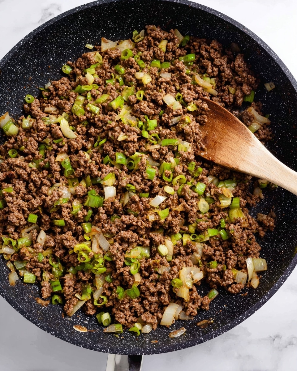 A close-up top view of a black speckled pan filled with ground meat cooked with chopped green onions and small pieces of white onion, creating a mix of brown, light green, and white colors. There is a wooden spoon resting inside the pan, partially covered by the cooked meat and vegetables. The pan is placed on a white marbled surface. photo taken with an iphone --ar 4:5 --v 7