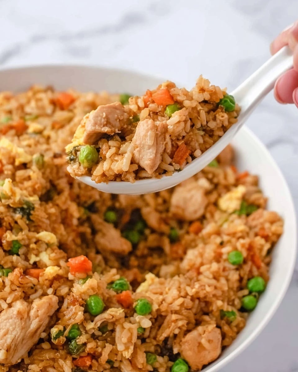 This image shows a close-up of a white spoon holding fried rice above a large white bowl filled with fried rice. The fried rice has grains of brown cooked rice mixed with small green peas, orange carrot cubes, and pieces of cooked chicken. The chicken pieces are light brown and mixed evenly throughout the rice. The background is a white marbled surface. A woman's hand is holding the spoon. The photo is bright and clear, showing the texture of the rice and ingredients well. photo taken with an iphone --ar 4:5 --v 7