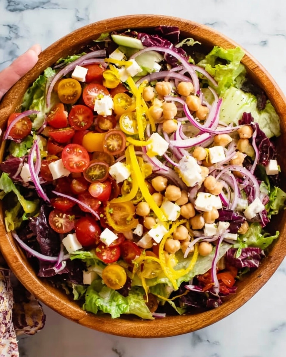 A wooden bowl filled with a colorful mixed salad sits on a white marbled surface. The bottom layer has green and purple leafy lettuce mixed together. On top, there are sliced cherry tomatoes, yellow pepper rings, chopped white cheese cubes, slices of red onion, and chickpeas spread evenly. The salad looks fresh with a mix of soft and crunchy textures from the different vegetables and cheese. A woman's hand is seen holding the edge of the bowl from the top corner. photo taken with an iphone --ar 4:5 --v 7