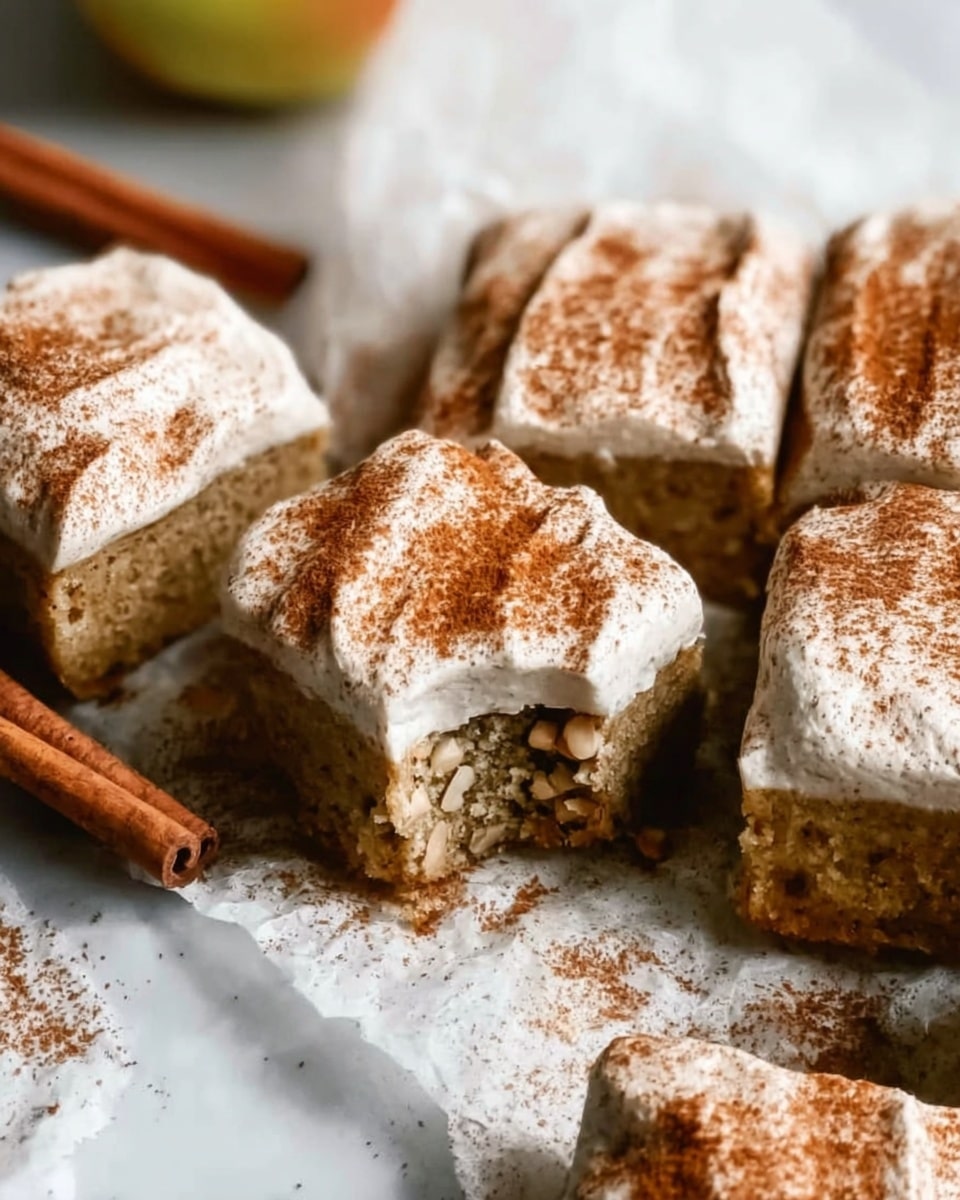 The image shows a close-up of several square pieces of light brown cake with a thick layer of white cream on top, each sprinkled with a fine dusting of cinnamon powder. One piece is turned over to reveal a moist, crumbly inside filled with small nut pieces, adding texture contrast. The cakes are closely placed on wrinkled parchment paper over a white marbled surface. In the background, cinnamon sticks and part of an apple are slightly out of focus, adding a natural touch to the setting. The light and shadows highlight the soft texture of the cream and the roughness of the cake beneath, making the dessert look inviting and fresh. Photo taken with an iphone --ar 4:5 --v 7