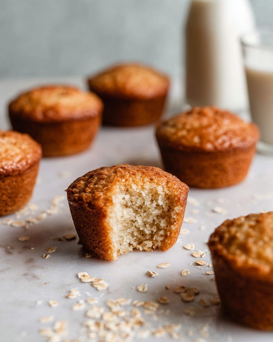 The image shows several small, round muffins with a golden-brown top and a light, soft crumb inside. The front muffin has a small piece bitten off, revealing its fluffy inside texture. The muffins are arranged loosely on a white marbled surface, with some scattered oats around them. In the background, there is a glass bottle filled with milk and a white cup, both slightly out of focus. The overall lighting is soft and natural, highlighting the warm color and texture of the muffins. photo taken with an iphone --ar 4:5 --v 7