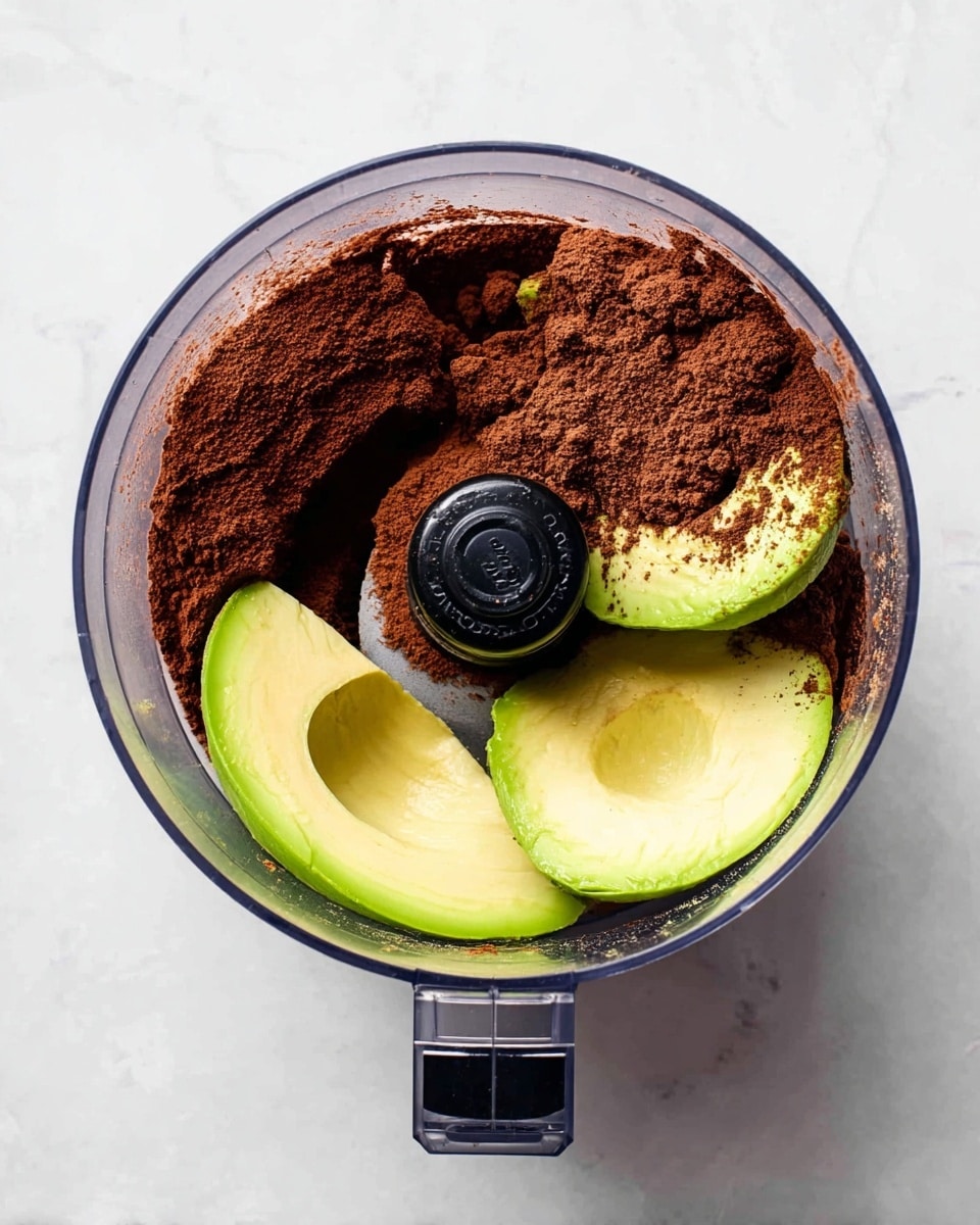 A clear round food processor bowl contains a mix of ingredients seen from above. The bottom layer is made of smooth bright green avocado halves with one half showing its brown seed cavity and the other half filled with a light beige liquid. On top of the avocado, there is a thick layer of dark brown cocoa powder spread unevenly. The food processor blade is in the center, black and round with cocoa powder resting on it. The whole setup is on a white marbled surface. photo taken with an iphone --ar 4:5 --v 7