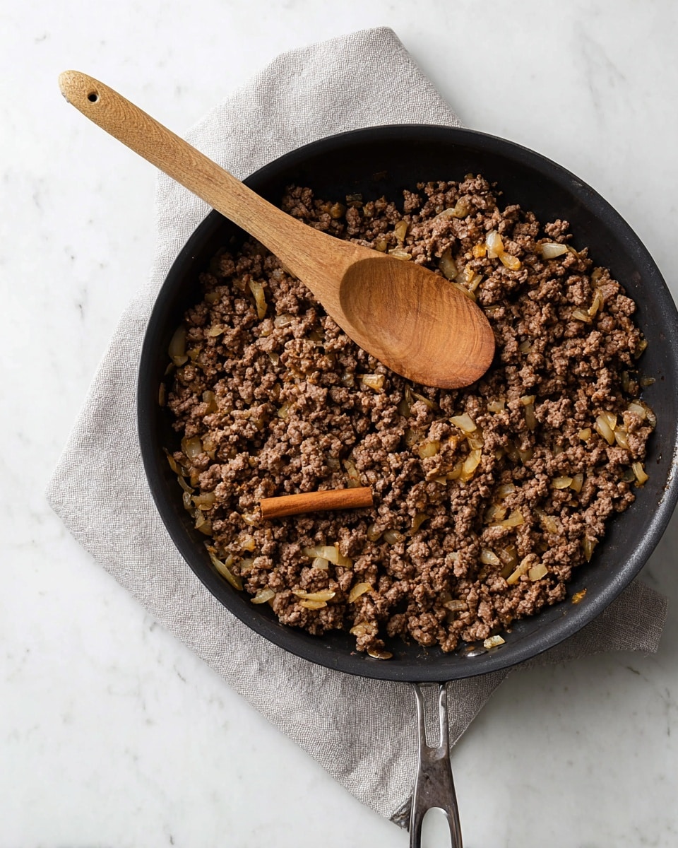 A large black pan filled with cooked ground meat mixed with small pieces of translucent cooked onions and a cinnamon stick in the center. A wooden spoon with a smooth texture rests on top of the meat, positioned diagonally from the bottom right to the middle. The pan is placed on a pale grey cloth over a white marbled surface, creating a simple and clean background. photo taken with an iphone --ar 4:5 --v 7