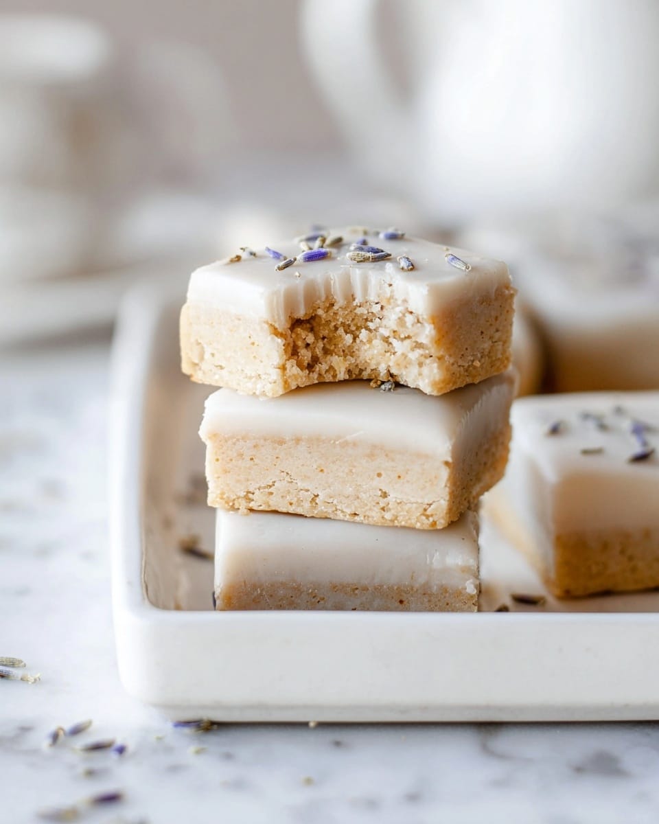 The image shows three stacked square bars with two layers each, placed on a white rectangular tray. The bottom layer of each bar is dense and crumbly with a light beige color, while the top layer is a smooth, glossy white icing. Small specks of lavender or similar tiny flowers are sprinkled on the icing, adding contrast with their purple tones. The top bar has a bite taken out from one corner, revealing the crumbly texture inside. The background is soft and blurred with a white marbled surface beneath the tray. Photo taken with an iphone --ar 4:5 --v 7