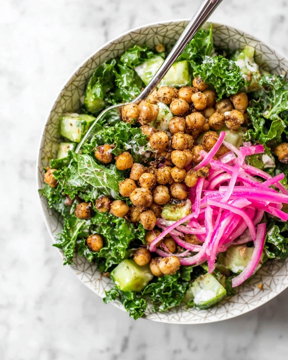 In a white bowl with a subtle geometric pattern, there is a mixed salad with three main layers. The base layer shows fresh, curly green kale leaves with a slightly rough texture. On top of the kale, there are light brown roasted chickpeas, seasoned and looking crunchy, spread evenly. Scattered among them are thin, bright pink rings of pickled onions, adding a pop of color and softness. Small chunks of green cucumber with smooth skin and light flesh are mixed throughout. The salad appears lightly dressed with a creamy, white sauce that adds slight gloss to the ingredients. A shiny silver fork is placed inside the bowl on the upper side. The bowl is sitting on a white marbled surface. Photo taken with an iphone --ar 4:5 --v 7