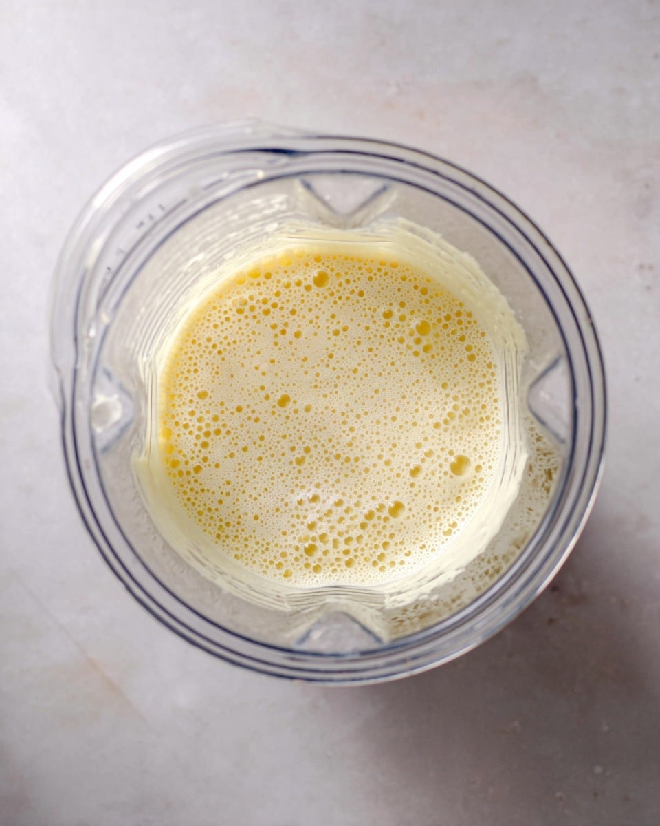 A clear blender cup filled halfway with a light yellow liquid that has many small bubbles on the surface, showing a smooth and slightly frothy texture. The blender is positioned at the center of a white marbled surface, with natural light highlighting the translucent container and the creamy mixture inside. Photo taken with an iphone --ar 4:5 --v 7
