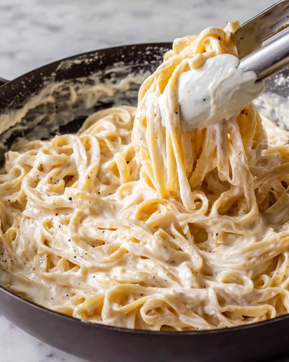 A close-up image of noodles tossed in a rich, creamy white sauce inside a dark pan. The noodles are thick and coated evenly with the sauce, which has a smooth texture with tiny black pepper specks. A pair of white tongs is lifting a portion of the noodles, showing the sauce clinging to each strand. The background features a white marbled surface. photo taken with an iphone --ar 4:5 --v 7