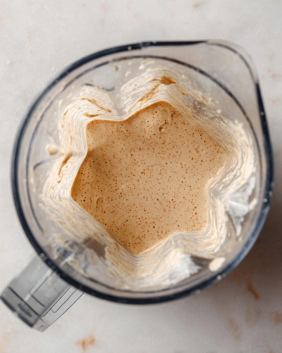 A clear blender jar filled with a smooth, creamy mixture that is light beige with small brown specks evenly spread throughout. The texture looks thick but slightly liquid, covering the bottom and a few higher spots inside the jar. The blender is placed on a white marbled surface. Photo taken with an iphone --ar 4:5 --v 7