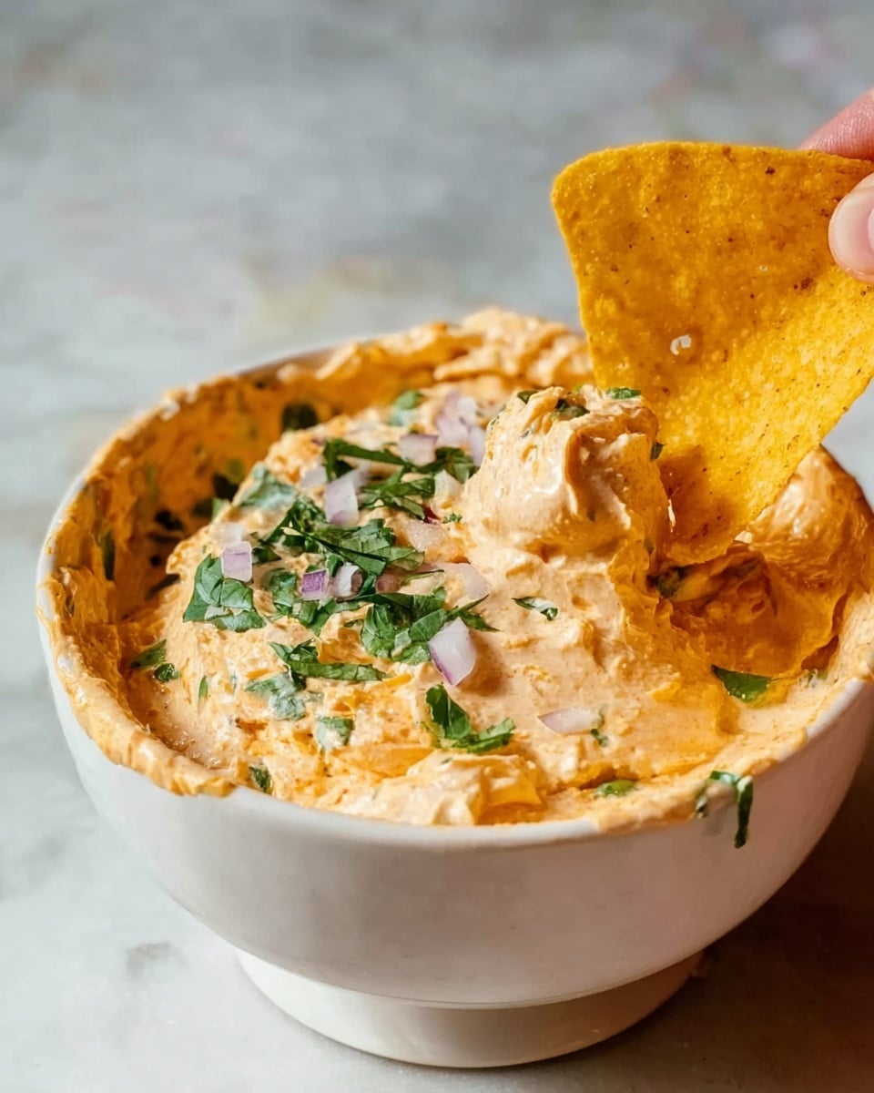 A white bowl filled with creamy, light orange dip that has a smooth texture and small green herb pieces mixed in. On top of the dip, there are chopped green herbs and small purple onion pieces scattered near the front edge. A large yellow tortilla chip is partially dipped in the creamy dip, held by a woman's hand, showing its rough texture and crunchy look. The background is a white marbled surface. photo taken with an iphone --ar 4:5 --v 7