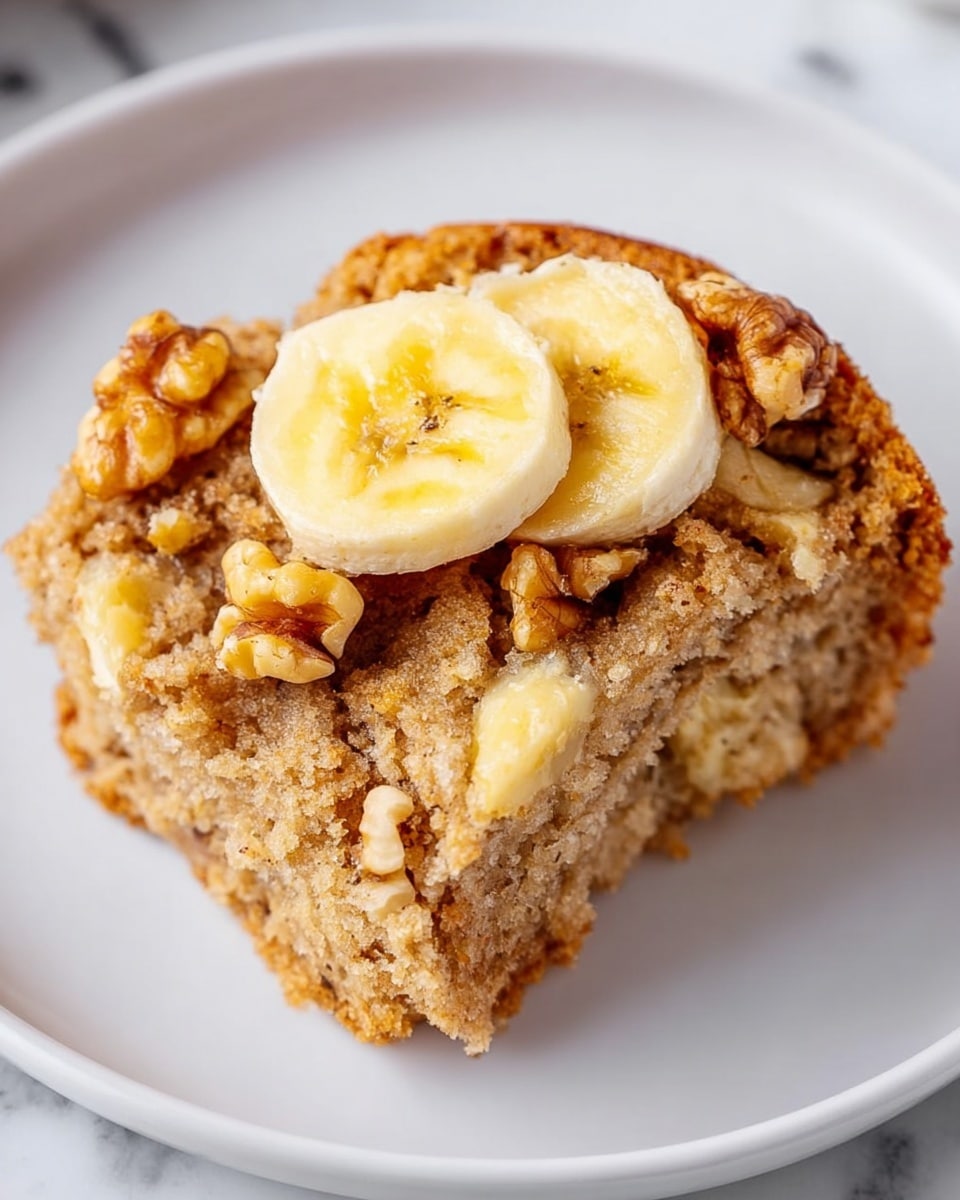 A close-up of a single serving of a moist, light brown cake slice on a white plate, with a soft, crumbly texture. On top of the cake, there are two thin slices of pale yellow banana and a generous amount of chunky light brown walnuts, some baked into the cake and some resting on top. The cake appears to have a slightly moist interior with pieces of banana visible, giving it a soft, dense look. The white plate rests on a white marbled surface. photo taken with an iphone --ar 4:5 --v 7