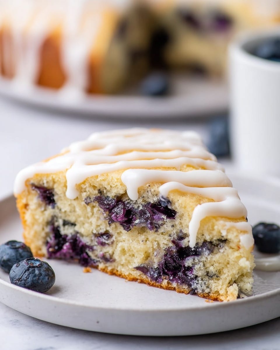A piece of soft, light golden cake with visible dark purple blueberries inside sits on a white plate. The cake has two layers: the bottom layer is the moist, fluffy cake embedded with juicy blueberries, and the top layer is drizzled with smooth white icing in loose stripes following the cake’s shape. The white marbled surface underneath the plate adds a clean, bright contrast. The background shows blurred pieces of similar cake. Photo taken with an iphone --ar 4:5 --v 7