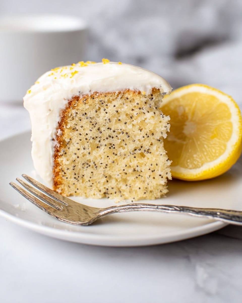A single slice of lemon poppy seed cake sits on a white plate with a shiny fork resting next to it. The cake has one thick layer that is light yellow with small black poppy seeds spread evenly inside. On top, there is a smooth layer of white frosting, slightly thick and creamy, covering the whole top surface. To the right of the cake slice, half a bright yellow lemon with visible juicy segments is placed upright. The background is a white marbled surface with soft lighting. Photo taken with an iphone --ar 4:5 --v 7