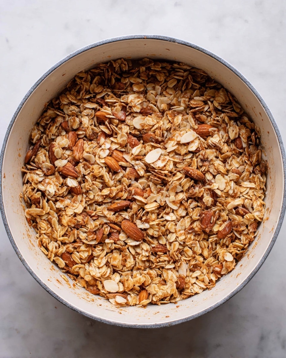 A white round pot filled with a single layer of crunchy granola mix. The granola has medium-sized rolled oats in light brown color, mixed with chopped almonds in a darker, rich brown shade. The pieces are uneven but well mixed, creating a textured surface with a slightly shiny, toasted look. The inside of the pot is cream-colored and clean except for some crumbs stuck lightly on the sides. The pot sits on a white marbled surface, giving a clean, bright background. photo taken with an iphone --ar 4:5 --v 7