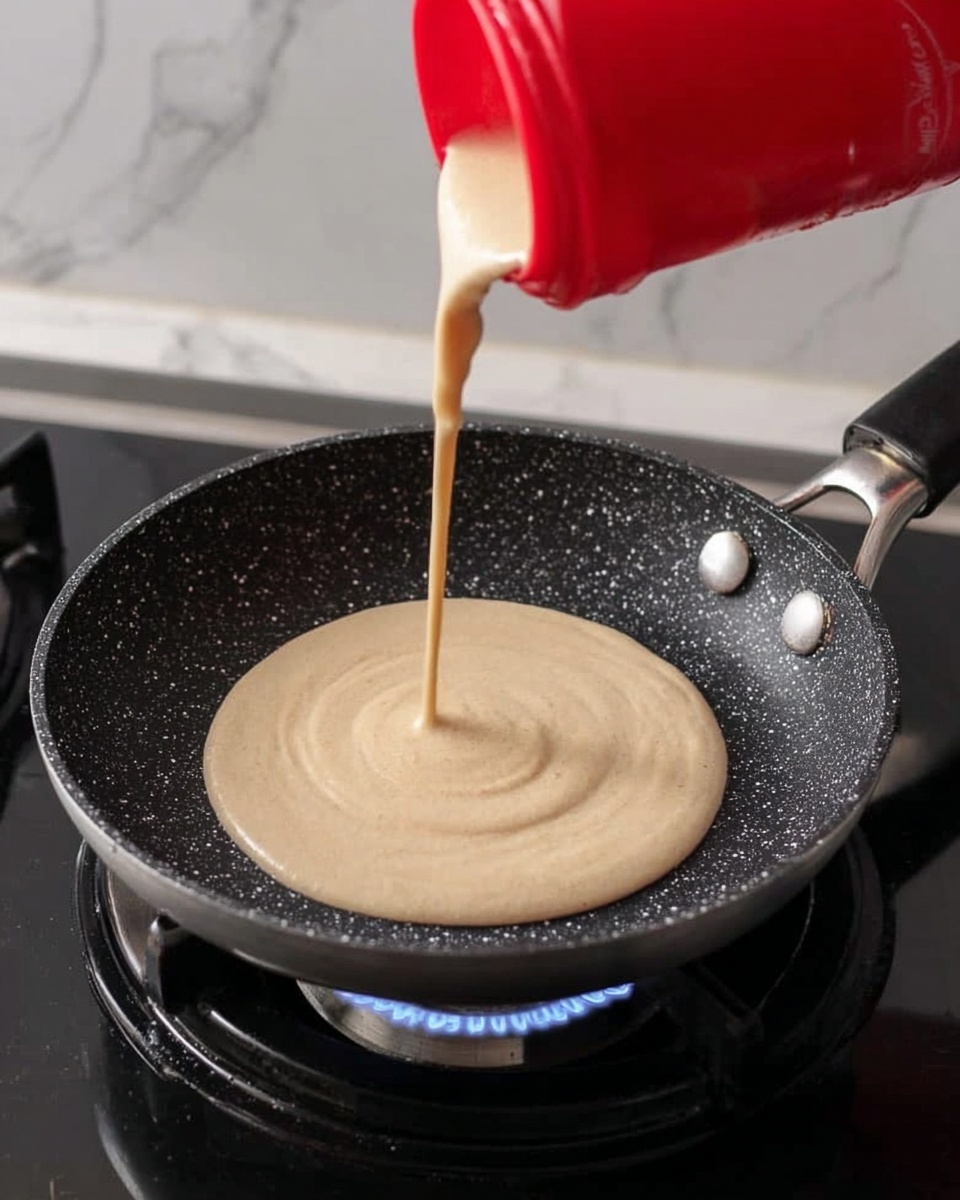 A close-up image shows light beige batter being poured from a red container into the center of a black speckled frying pan with a silver handle, positioned on a gas stove burner with a gentle blue flame below. The pouring batter forms a smooth, circular layer that is slightly thicker in the middle, creating a small mound in the pan. The background features a white marbled texture, enhancing the focus on the pan and the batter. photo taken with an iphone --ar 4:5 --v 7