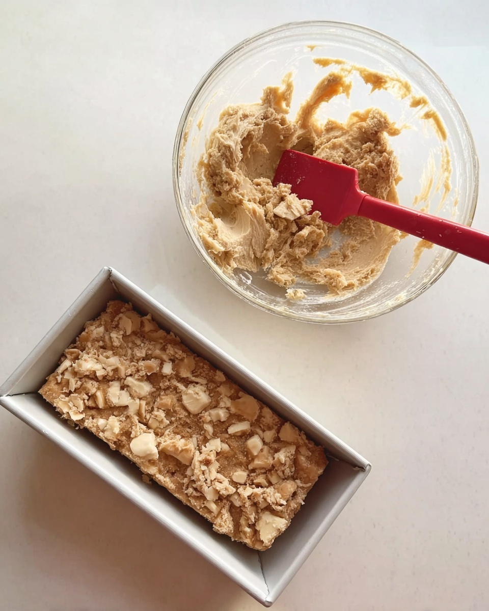 A clear glass bowl holding a thick mixture of thick, uneven pieces of light beige and cream ingredients mixed together, with a red spatula resting inside. Next to it, a white metal loaf pan on a white marbled surface, filled with a chunky mixture of light brown and cream pieces spread evenly. Photo taken with an iphone --ar 4:5 --v 7