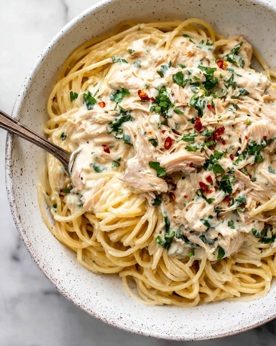 The image shows a white speckled bowl filled with creamy spaghetti. The bottom layer is light yellow cooked spaghetti, topped with a thick pale cream-colored sauce mixed with shredded light brown chicken pieces. Bright green chopped herbs and small red chili flakes are scattered on top, adding color contrast. A silver fork with a rustic handle is twirling some spaghetti at the center of the bowl. The bowl sits on a white marbled surface. Photo taken with an iphone --ar 4:5 --v 7