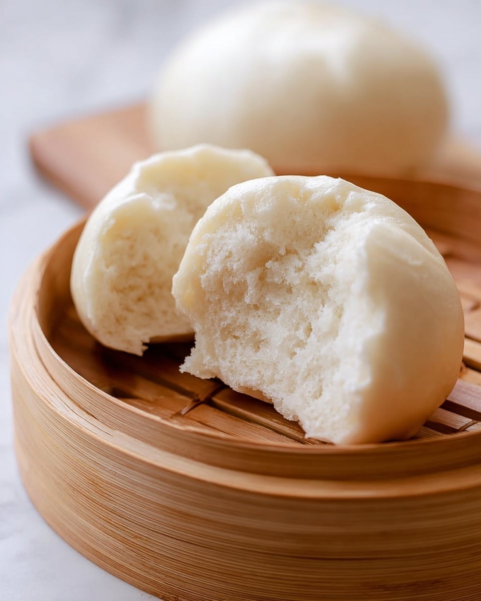 The image shows a close-up of a white soft steamed bun that is torn open to show its fluffy and light inside texture. The bun is round with a smooth surface and placed on the base of a round bamboo steamer. The steamer has a warm brown color with its wooden slats visible underneath the bun. Another whole steamed bun sits blurred in the background inside the steamer. The background is a white marbled texture, creating a clean and bright setting. Photo taken with an iphone --ar 4:5 --v 7