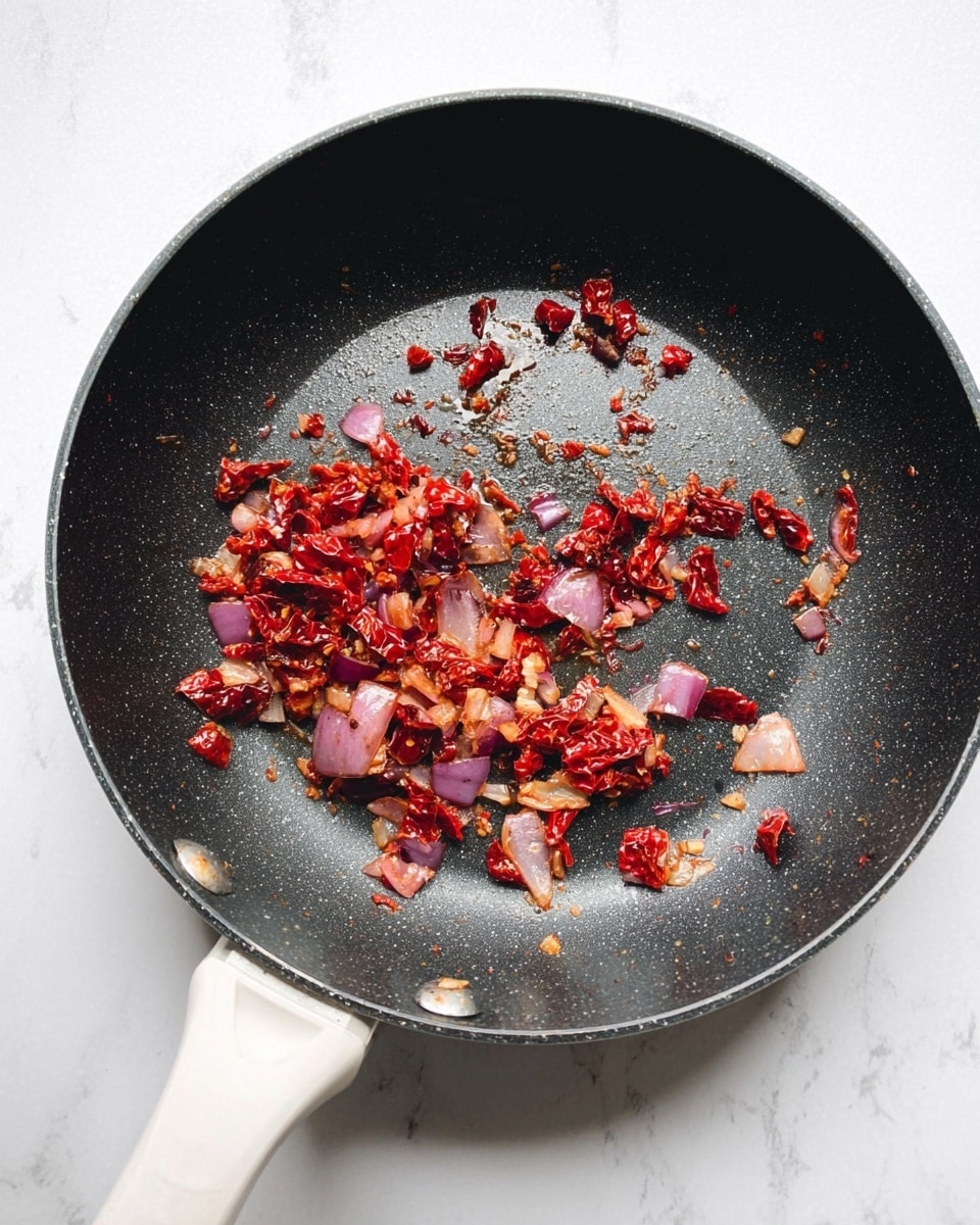 A black frying pan with a white handle contains small pieces of cooked red onion and sun-dried tomatoes scattered across its center. The vegetables look soft with a mix of deep red from the tomatoes and light purple from the onions, showing a slightly oily texture as if sautéed. The pan is placed on a white marbled surface. photo taken with an iphone --ar 4:5 --v 7