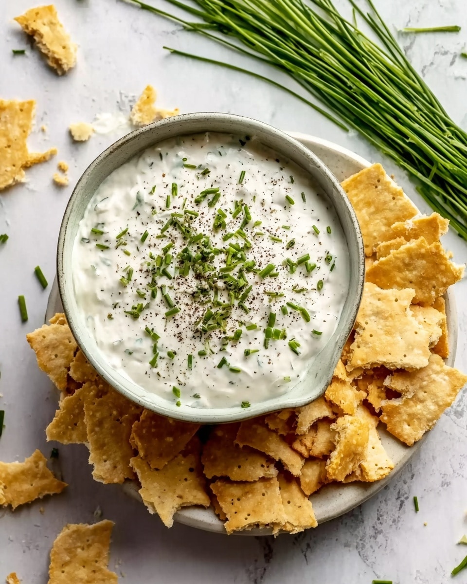 A white bowl filled with a creamy white dip sprinkled generously with chopped green herbs and black pepper on top, placed on a white plate. Around the bowl, there are broken pieces of light golden crackers scattered on the white marbled surface. To the side, there is a bunch of fresh green chives adding a fresh touch to the scene. The photo taken with an iphone --ar 4:5 --v 7