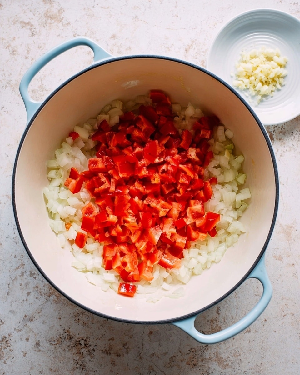 A white cooking pot with light blue handles sits on a white marbled surface, filled with two layers of chopped vegetables. The bottom layer is small white onion pieces covering most of the pot base, and on top is a layer of red bell pepper cubes centered more in the middle, showing vibrant red hues with a slightly shiny texture. To the top right of the pot, a small white plate holds a small pile of finely chopped garlic. The image is clear and bright, showing fresh ingredients ready to cook, photo taken with an iphone --ar 4:5 --v 7