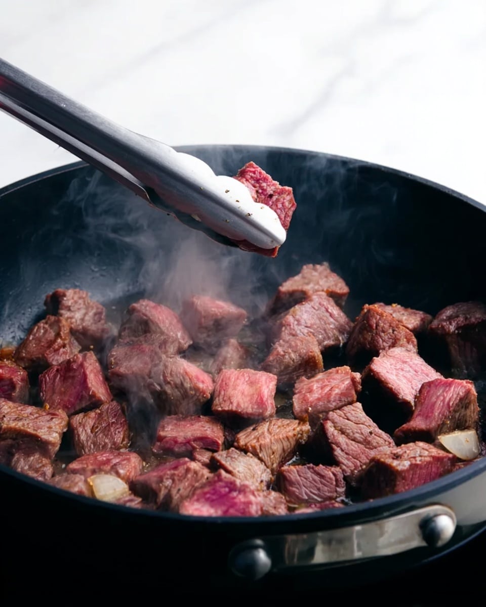 A close-up view of a black pan filled with small chunks of beef being cooked, with some pieces browned on the edges and others still pink in the middle. A woman's hand is holding silver tongs, lifting one piece of meat above the pan. Steam rises from the pan, showing that the meat is hot. The background is a white marbled texture photo taken with an iphone --ar 4:5 --v 7