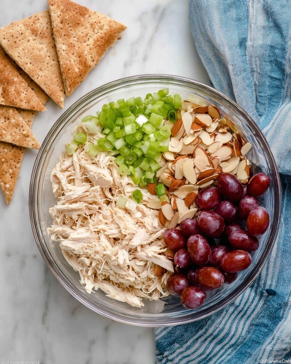 A clear glass bowl contains four separate layers of ingredients, arranged side by side. The bottom right has shredded light beige chicken, the bottom left shows a pile of thinly sliced almond pieces in light brown and cream colors, the top left holds finely chopped green onions with bright green and white colors, and the top right is filled with halved red grapes showing their dark red skin and light green interiors. To the left of the bowl, there are triangular pieces of brown pita bread slightly stacked, and to the right, a blue and white striped cloth rests on the white marbled surface beneath the bowl. Photo taken with an iphone --ar 4:5 --v 7