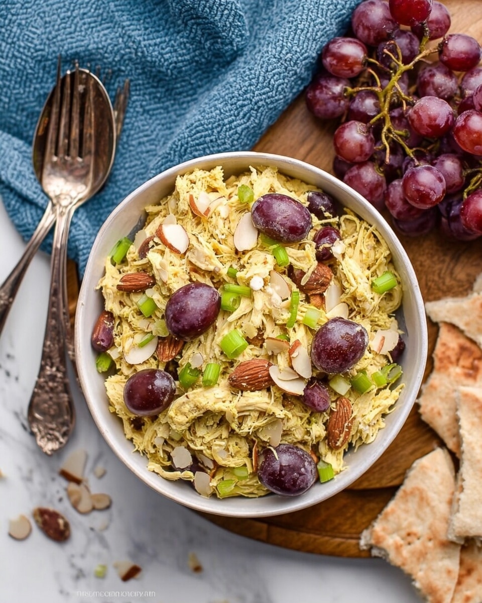 A white bowl filled with shredded yellow chicken mixed with sliced purple grapes, thin green onion slices, and small almond slivers scattered on top. The bowl sits on a wooden board next to a blue textured cloth with two vintage forks resting on it. A bunch of red grapes lies on the wooden board near the bowl, and some folded pita bread pieces are placed on the white marbled surface next to the bowl. Photo taken with an iphone --ar 4:5 --v 7