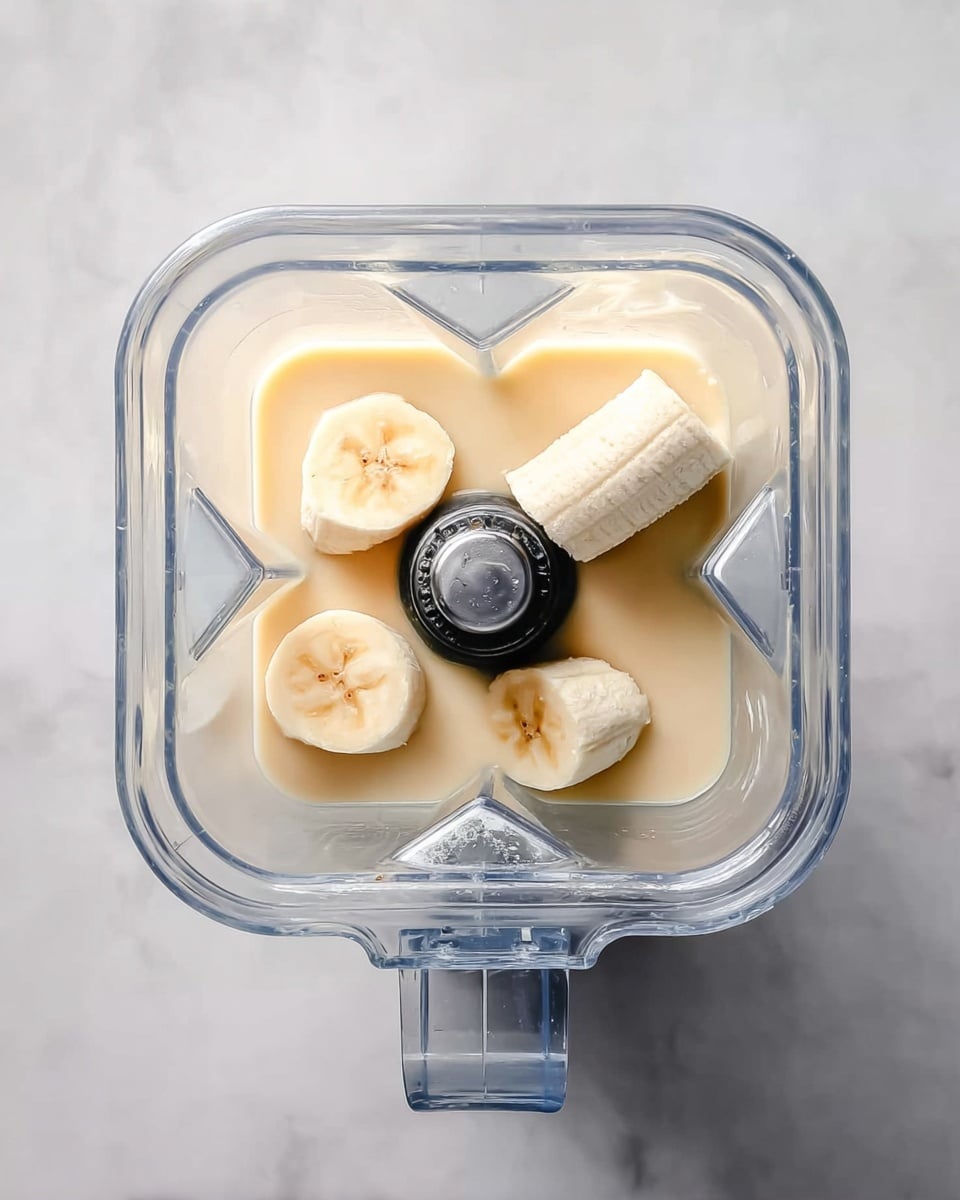 Top view of a square blender pitcher showing three pieces of peeled banana placed on a smooth light beige liquid inside. The blender blades are sharp and in the center beneath the bananas. The pitcher is clear and sits on a white marbled surface. Photo taken with an iphone --ar 4:5 --v 7