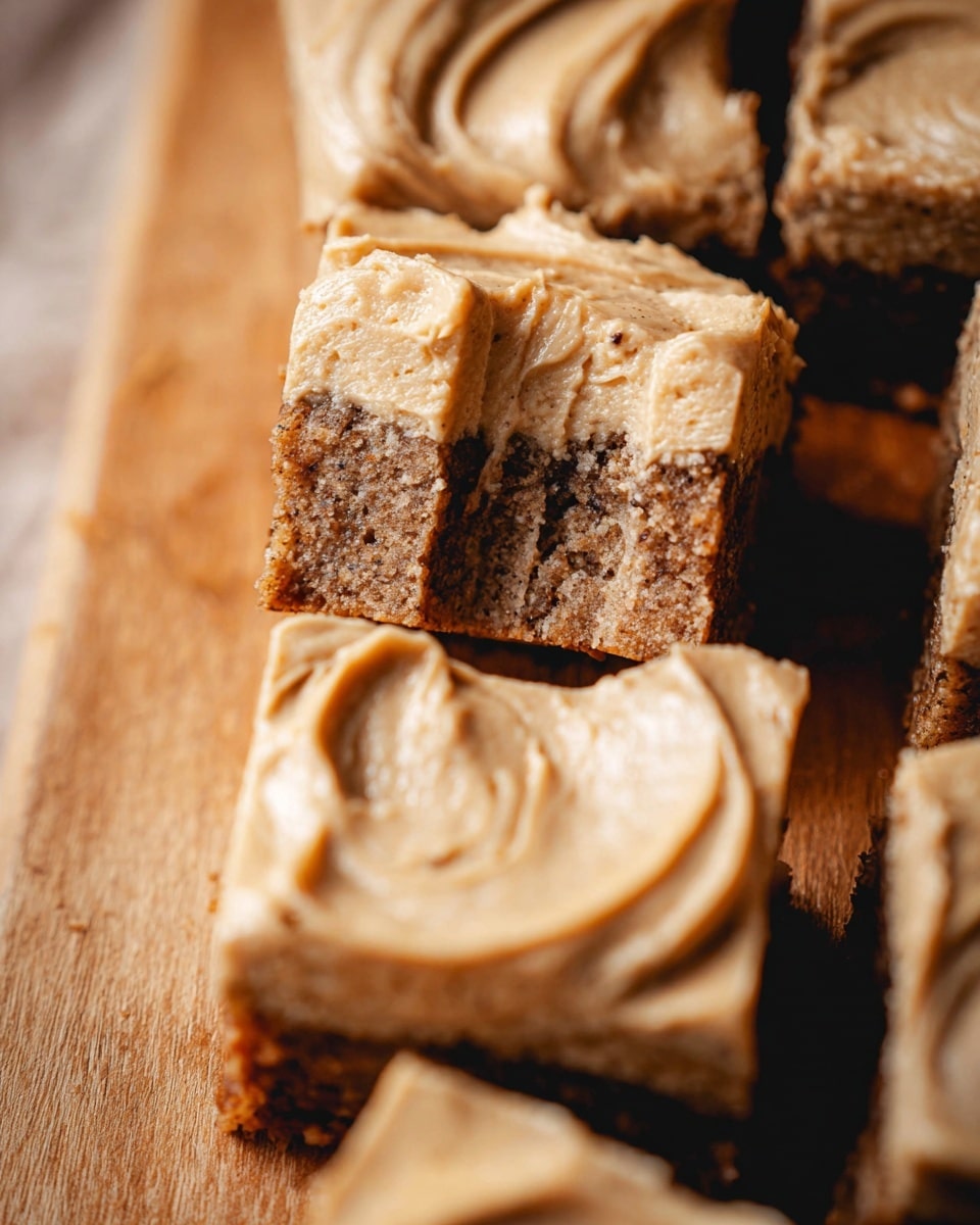 The image shows several square-shaped pieces of cake layered on a wooden board with a light brown color tone. Each piece has two layers: the bottom layer is a dense cake with a slightly crumbly texture and a darker brown color with hints of nutty or grainy bits, while the top layer is a smooth, creamy frosting in a light caramel or tan color spread evenly with soft, swirling patterns. One piece is broken in the middle, revealing the inside texture of the cake layer. The background is softly blurred, focusing fully on the cake pieces. photo taken with an iphone --ar 4:5 --v 7