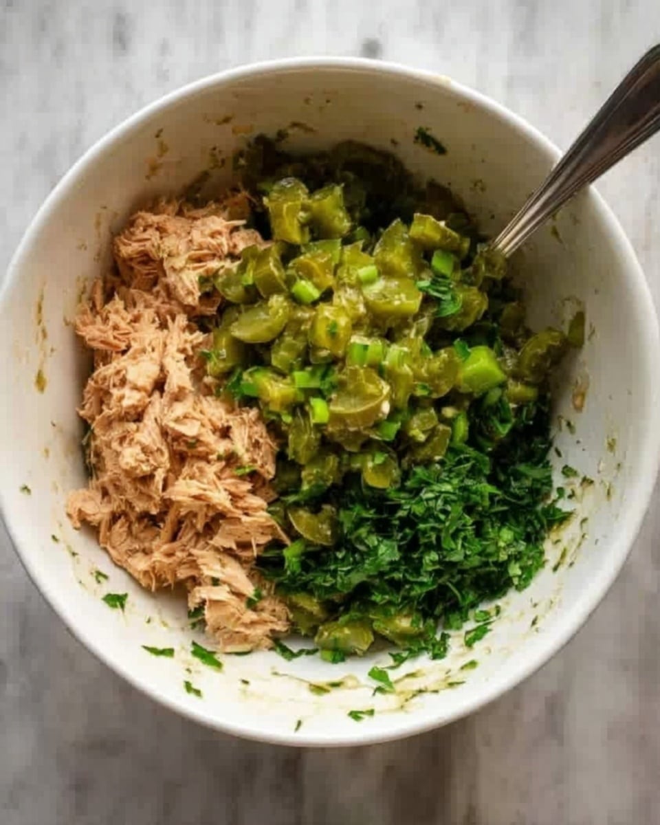 A white bowl sits on a white marbled surface, filled with three mixed ingredients before being stirred. On the left side of the bowl, there is a light brown, shredded mixture resembling tuna. On the right side, bright green chopped pickles cover part of the bowl. Below the pickles, fresh chopped herbs add a darker green color to the mix. A metal spoon rests inside the bowl on the right side, partially sinking into the ingredients. The bowl shows light marks and smudges from mixing. photo taken with an iphone --ar 4:5 --v 7