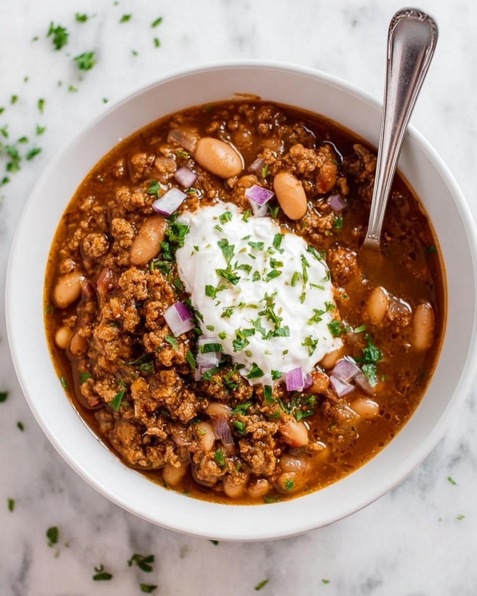A white bowl filled with a rich brown stew that has pieces of cooked ground meat and white beans throughout. On top, there is a dollop of white sour cream sprinkled with small green chopped herbs. There are small chunks of purple onion and more green herbs scattered on the surface of the stew. A silver spoon is placed inside the bowl, resting on the stew. The bowl is placed on a white marbled surface with a few small green herb bits around. Photo taken with an iphone --ar 4:5 --v 7
