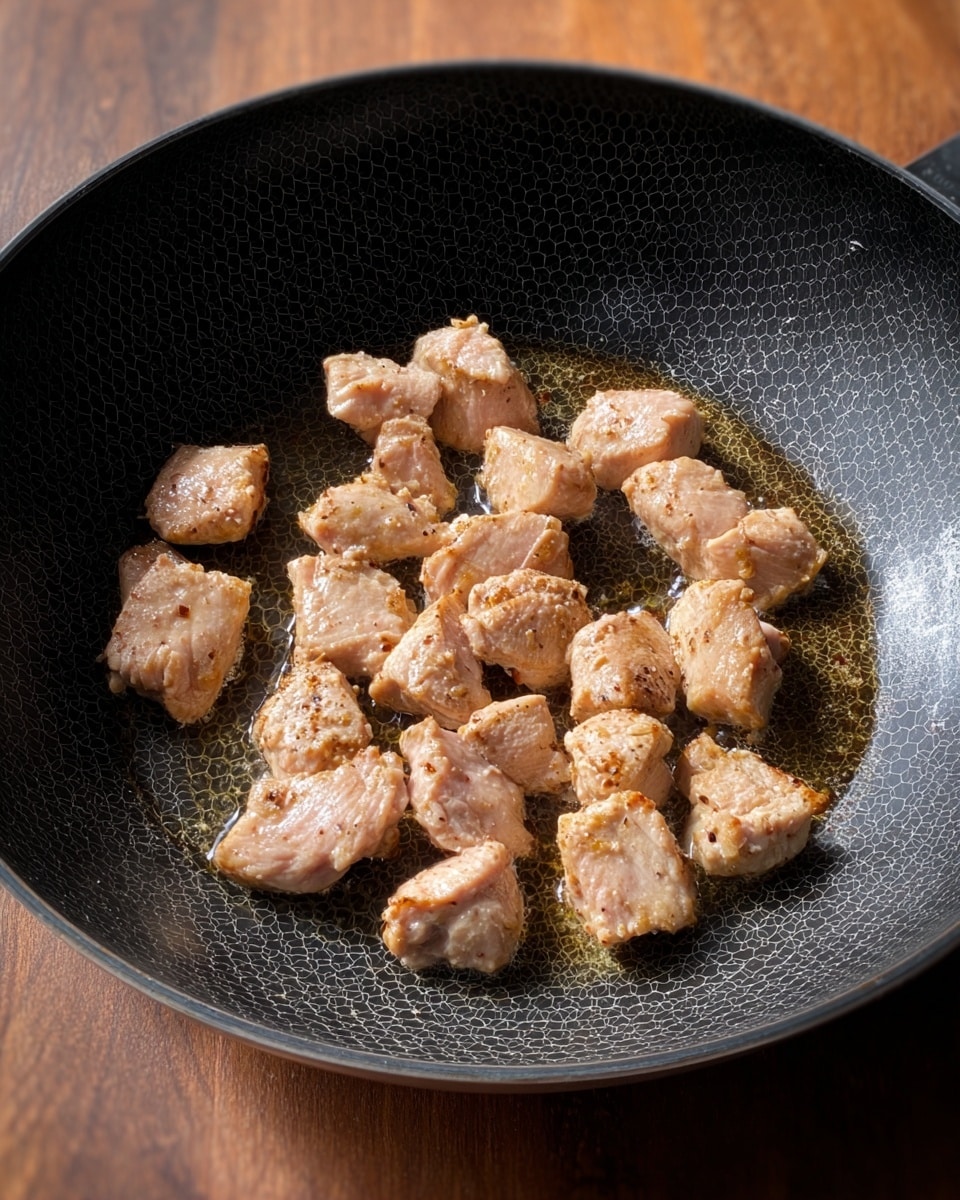 Inside a black frying pan with a hexagon pattern, small pieces of light pink chicken are cooking in oil. The chicken pieces are scattered in the center, each showing a slightly browned and textured surface from the cooking process. The pan sits on a wooden surface. The light in the image highlights the wet shininess of the oil and the slight crispiness forming on the chicken edges. photo taken with an iphone --ar 4:5 --v 7