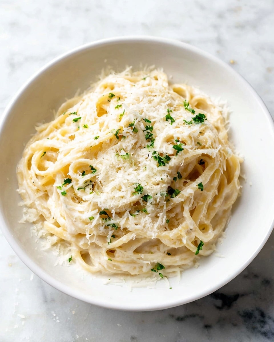A white bowl filled with creamy pasta showing long, thick noodles coated in a smooth white sauce. On top, a generous layer of finely grated white cheese covers the pasta, with small green herb pieces scattered lightly throughout. The bowl sits on a white marbled surface with a few cheese shreds around it, giving a fresh and simple look. photo taken with an iphone --ar 4:5 --v 7