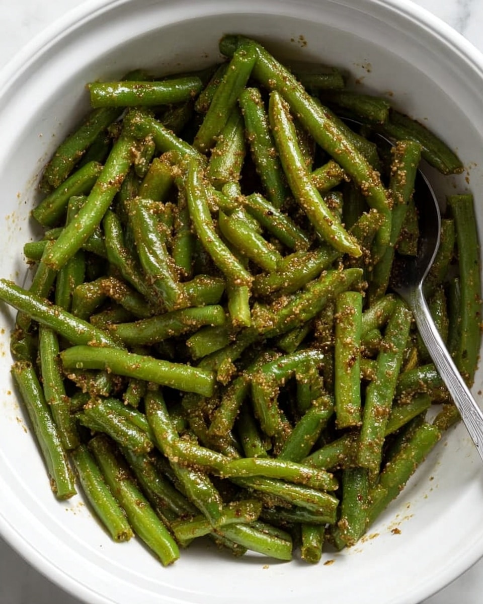 A white bowl filled with a generous layer of cooked green beans, all cut into similar lengths. The green beans have a light coating of a grainy, brown seasoning that is evenly spread across them, giving a textured appearance. A silver spoon is at the right edge inside the bowl, partially covered by the green beans. The bowl is placed on a white marbled surface. photo taken with an iphone --ar 4:5 --v 7