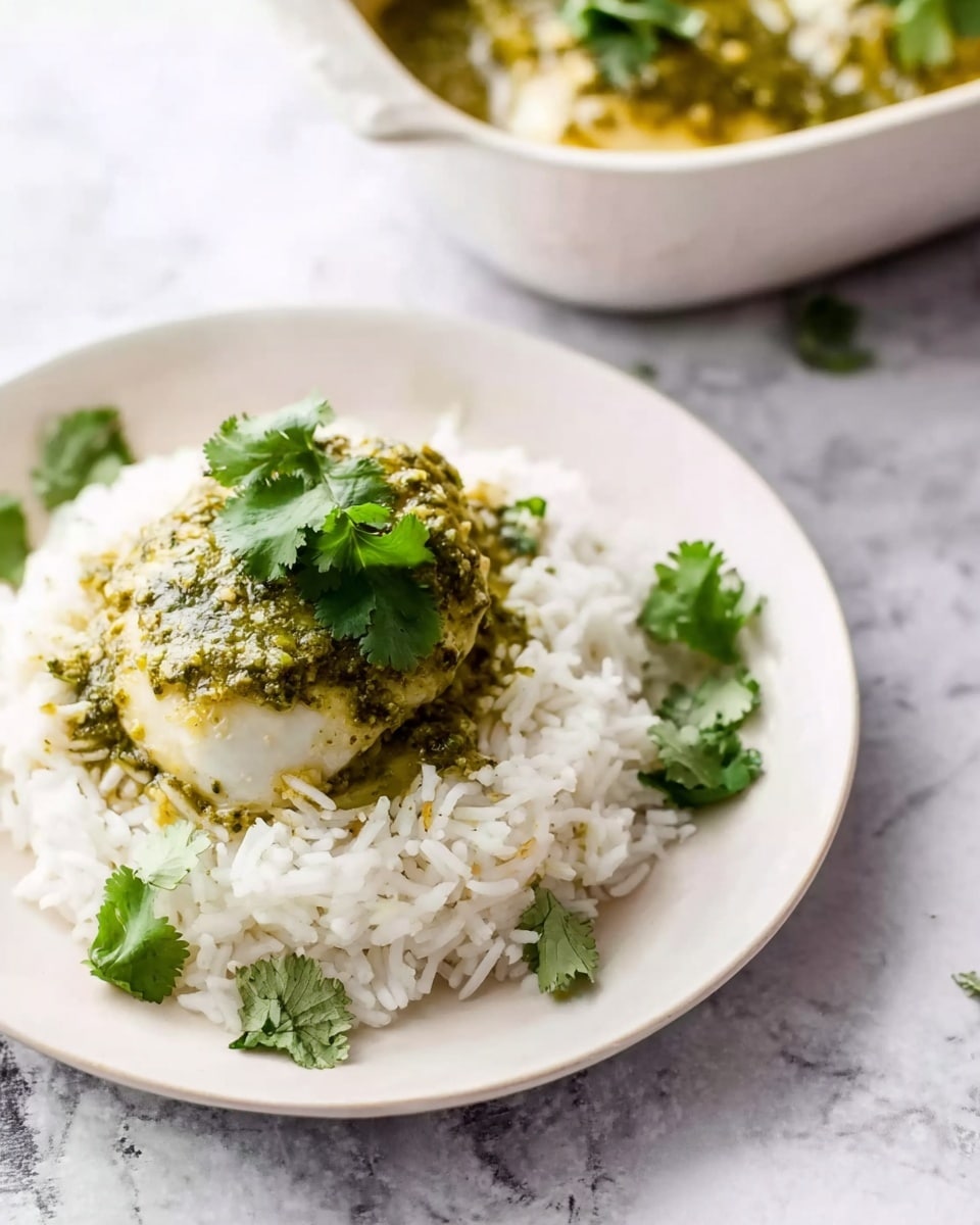 The image shows a white plate with a small portion of fluffy white rice forming a round base. On top of the rice, there is a cooked chicken piece covered with a green sauce that has a slightly chunky texture. The chicken is topped with fresh green cilantro leaves for garnish. In the background, a white bowl filled with more chicken and green sauce is placed on a white marbled surface. A woman's finger is gently touching the bowl's side. Photo taken with an iphone --ar 4:5 --v 7