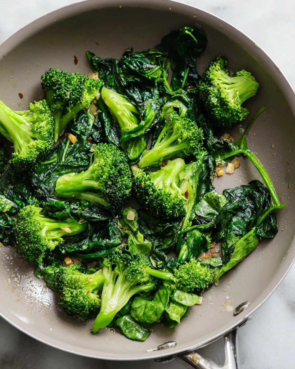 This image shows cooked broccoli florets mixed with wilted dark green spinach leaves in a light gray frying pan. The broccoli pieces are bright green with some brown spots where they are slightly cooked or roasted, and small bits of garlic or seasoning are scattered around. The spinach leaves are dark green, soft, and slightly shiny, laying around and under the broccoli. The pan has a smooth texture, with small droplets of oil or moisture visible. The background is a white marbled texture. photo taken with an iphone --ar 4:5 --v 7