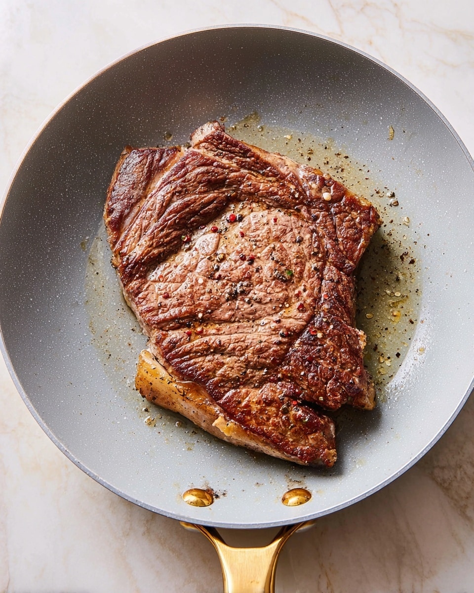 A large, browned steak with a textured surface showing a mix of light and dark brown cooked areas sits in the center of a light gray frying pan. The steak’s surface has small, scattered black and red pepper specks, and it is glistening slightly with cooking oil around its edges. The pan has a shiny golden handle extending downward, resting on a white marbled surface. The steak fills most of the pan’s base with some small oil droplets visible around it, and the pan’s rim is visible at the edges of the image. Photo taken with an iphone --ar 4:5 --v 7