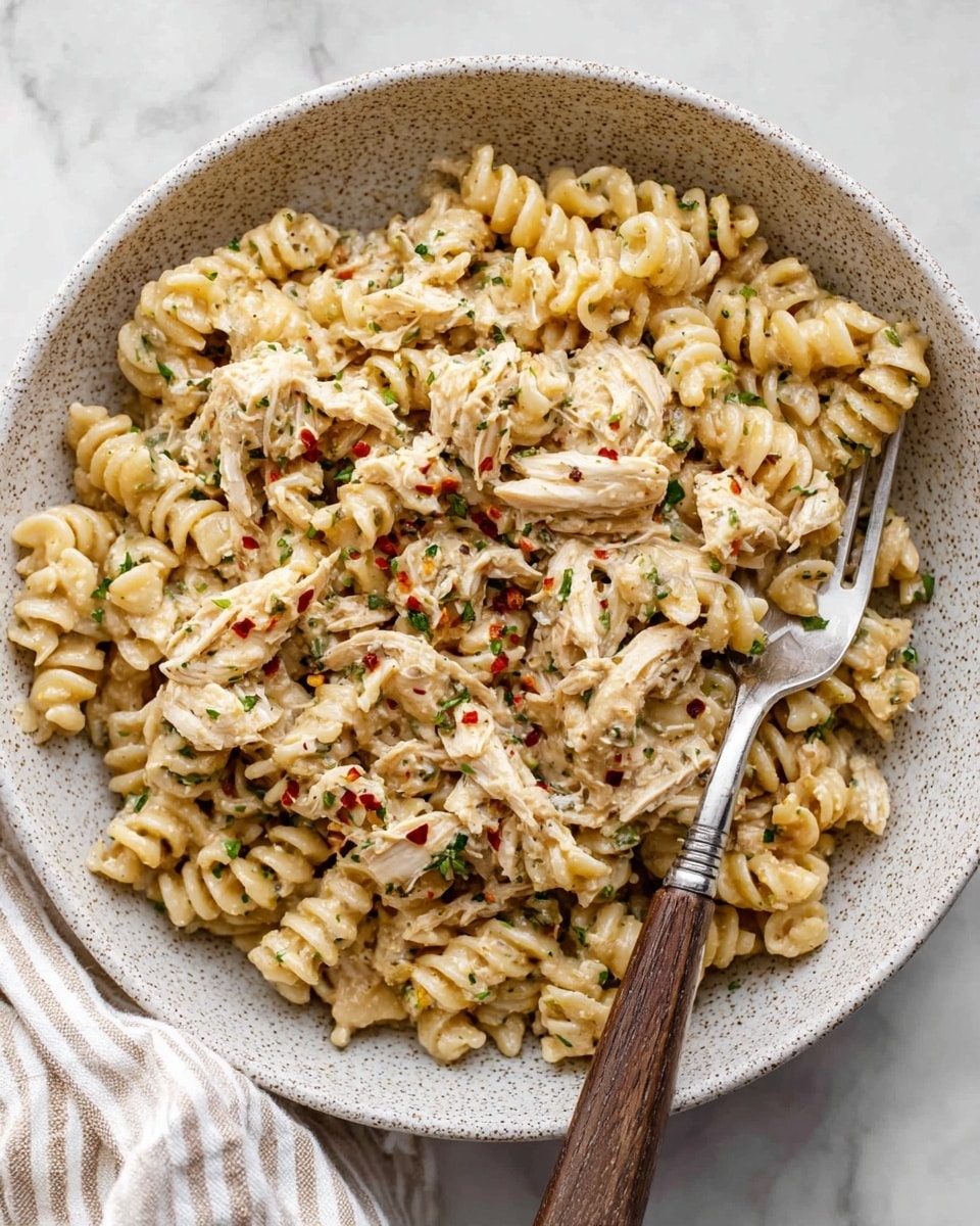 A white speckled bowl holds a creamy pasta dish made with short, twisted rotini noodles mixed with shredded chicken pieces. The pasta is coated in a thick, pale beige sauce with visible small green herbs and red chili flakes sprinkled on top. The texture looks rich and creamy, with the chicken and noodles blending evenly throughout. A silver fork with a wooden handle rests inside the bowl, slightly buried in the pasta. The bowl sits on a white marbled surface with a white and beige striped cloth nearby. Photo taken with an iphone --ar 4:5 --v 7
