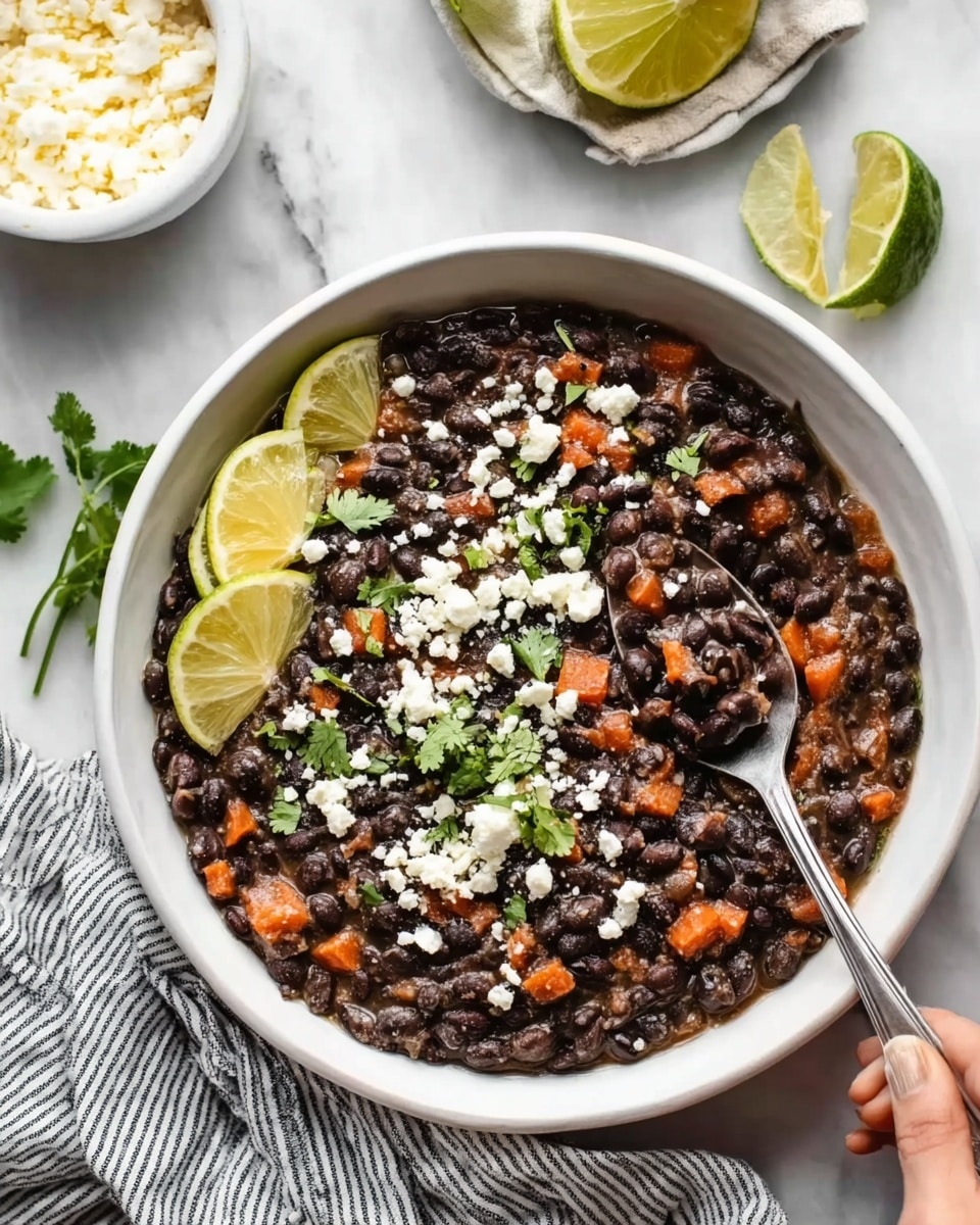 The image shows a white bowl filled with cooked black beans mixed with small orange carrot pieces, garnished with crumbled white cheese and fresh green cilantro leaves on top. Two thin lemon wedges are placed on the left edge of the bowl next to a silver spoon scooping some beans. The bowl sits on a white marbled surface next to a white bowl of cheese and a small white bowl with lime wedges. A woman's hand is holding the spoon inside the bowl. The setting includes a striped cloth underneath the bowl. photo taken with an iphone --ar 4:5 --v 7