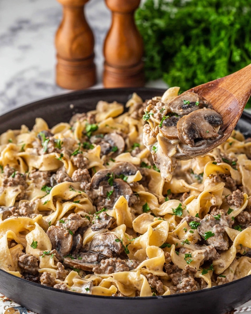 A black skillet is filled with creamy pasta made of wide egg noodles mixed with browned ground meat and sliced brown mushrooms, all coated in a light beige sauce. Small green parsley leaves are sprinkled across the dish for color. A wooden spoon is lifting some pasta, mushrooms, and meat from the skillet. The background shows a white marbled surface with a blurred bunch of green parsley and wooden pepper mills. Photo taken with an iphone --ar 4:5 --v 7