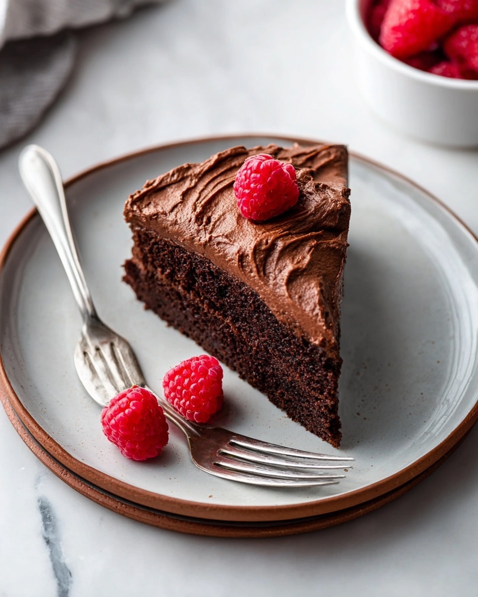 A slice of chocolate cake with one thick layer lies centered on a round white plate with a brown rim. The cake is topped with thick swirls of smooth, rich chocolate frosting that cover the entire top layer. A single bright red raspberry sits on top of the frosting in the center. Next to the cake on the plate are two more red raspberries, showing their bumpy texture. A silver fork lies at the bottom left of the plate touching the cake slice. The plate rests on a white marbled surface, and part of a white bowl with more red raspberries is visible in the top right corner. photo taken with an iphone --ar 4:5 --v 7